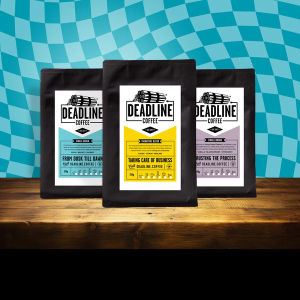 Three 'Deadline Coffee' packages on a wooden surface with a blue checkered background